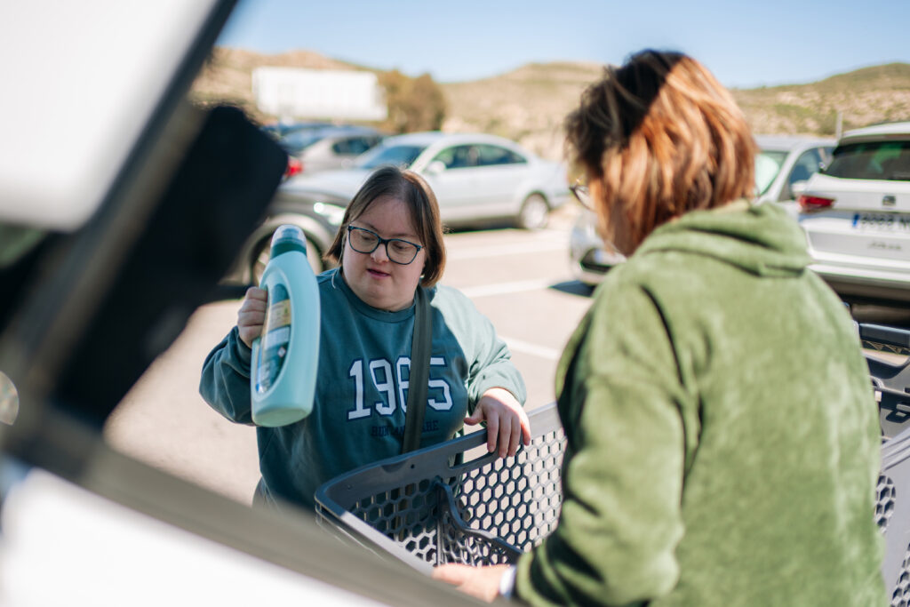 A woman with Down syndrome places a large bottle into a shopping cart in a parking lot while another woman assists nearby.
