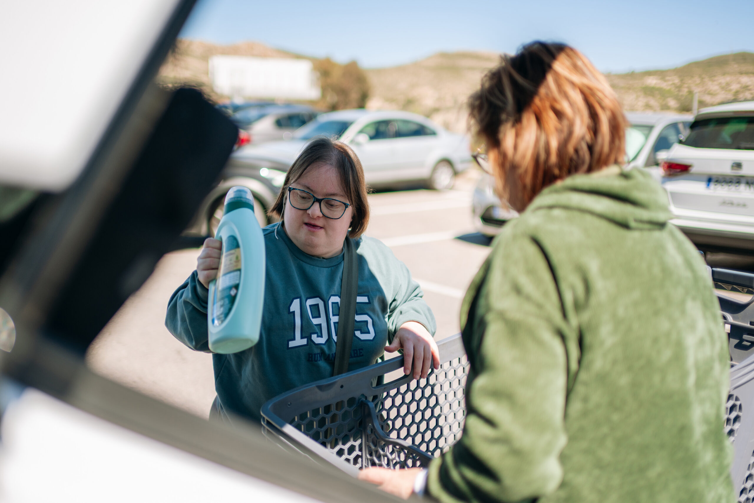 A woman with Down syndrome places a large bottle into a shopping cart in a parking lot while another woman assists nearby.