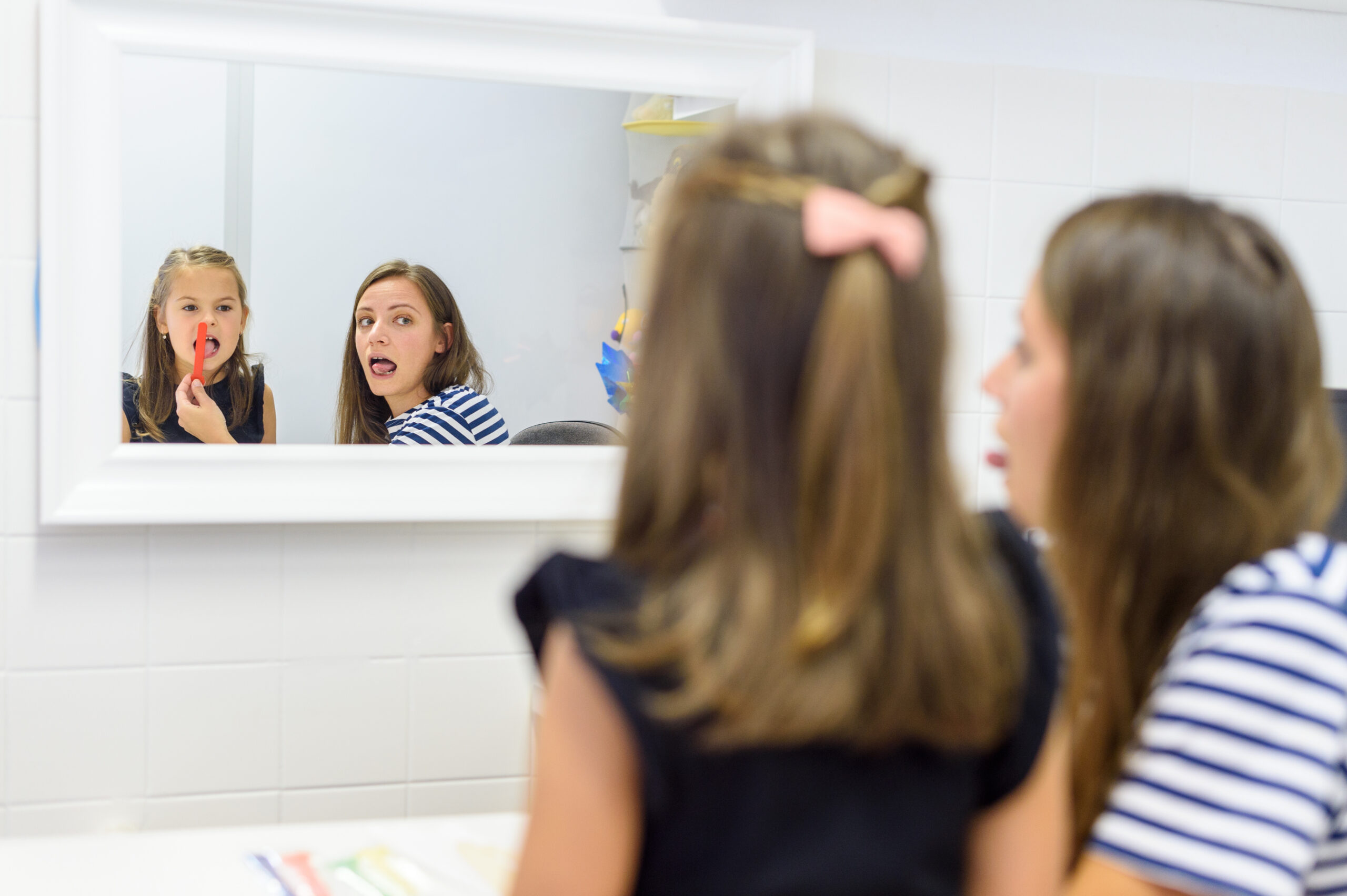 A speech therapist works with a young girl in front of a mirror during a therapy session.