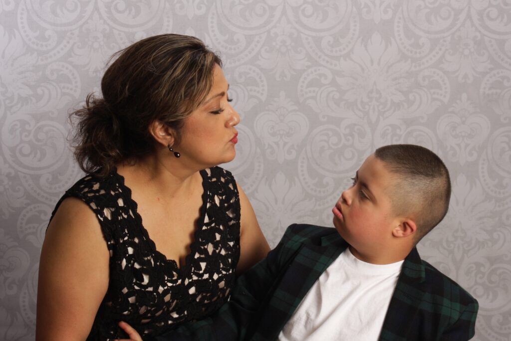 A mother and her son sit together, facing each other and making eye contact. She leans toward him with a playful expression as he looks up at her.