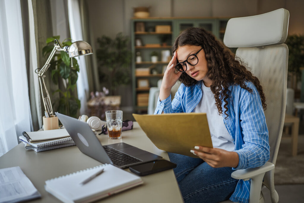 Stressed young woman at desk with laptop reviewing paperwork